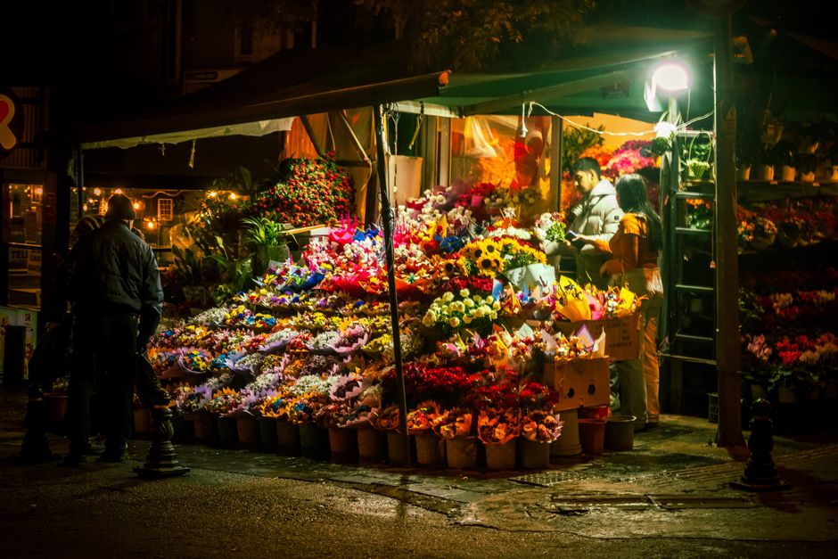 At a flower stall on Adelaide Road Swiss Cottage during the evening, an array of fresh flower bouquets and arrangements are displayed under warm lighting. The flowers feature vibrant colours, includin