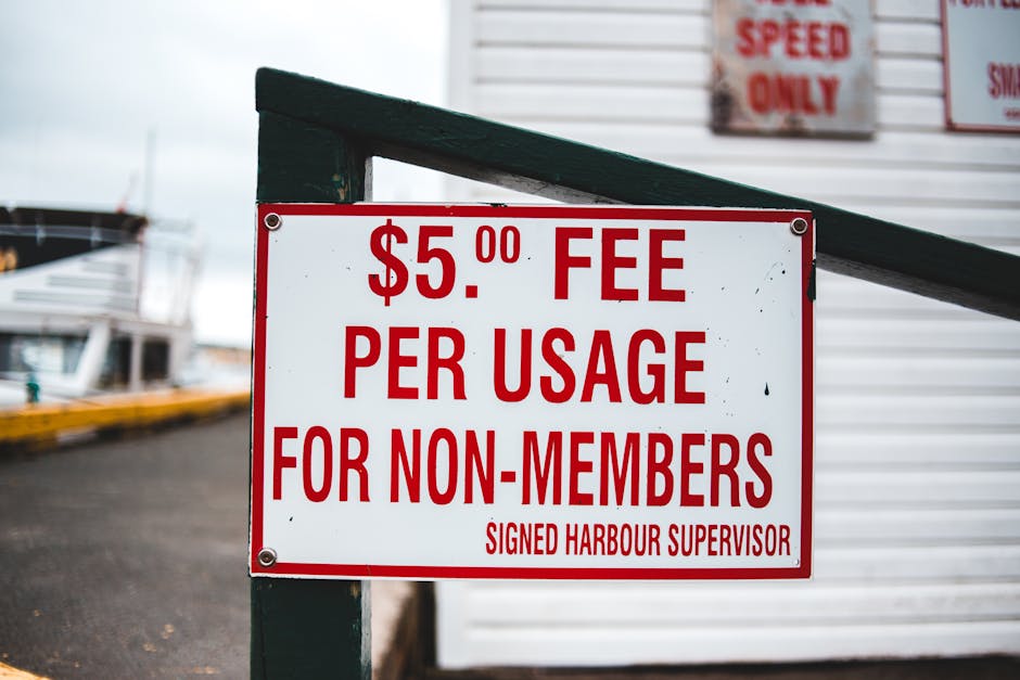 A rectangular white sign with bold red lettering displaying a fee of $5.00 per usage for non-members, attached to a dark green metal railing at a harbor. In the background, there are boats docked alon